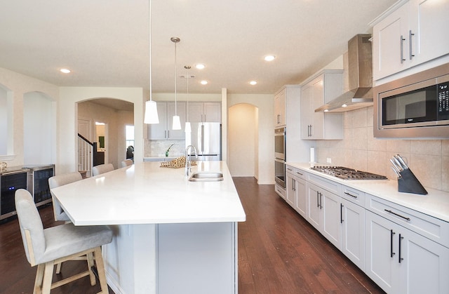 kitchen featuring pendant lighting, a center island with sink, sink, wall chimney exhaust hood, and appliances with stainless steel finishes