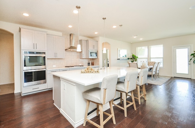 kitchen featuring white cabinets, hanging light fixtures, wall chimney exhaust hood, an island with sink, and appliances with stainless steel finishes