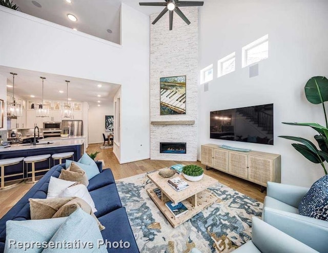 living room featuring light wood-type flooring, a towering ceiling, ceiling fan, sink, and a stone fireplace