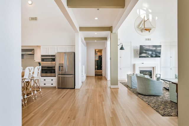 kitchen featuring white cabinetry, beamed ceiling, a towering ceiling, appliances with stainless steel finishes, and light wood-type flooring