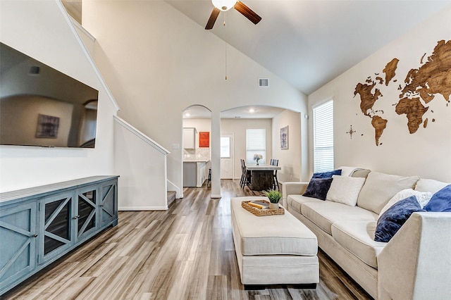 living room with arched walkways, visible vents, light wood-style floors, a ceiling fan, and stairs