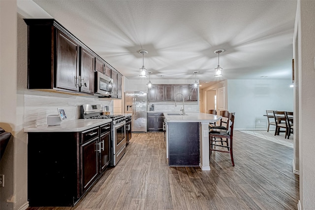 kitchen with hanging light fixtures, wood-type flooring, a kitchen bar, a center island with sink, and appliances with stainless steel finishes