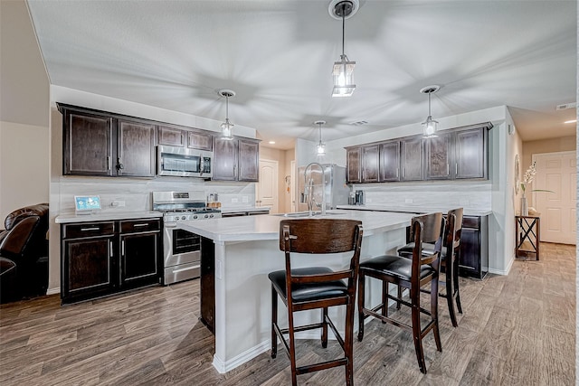 kitchen featuring a center island with sink, hanging light fixtures, tasteful backsplash, dark brown cabinetry, and stainless steel appliances