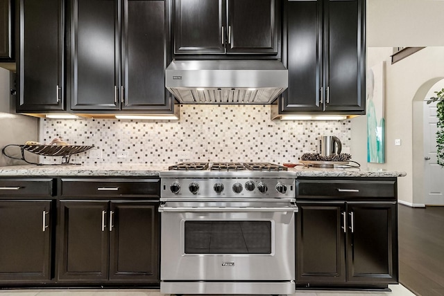 kitchen with decorative backsplash, high end stainless steel range, light stone counters, and wall chimney range hood