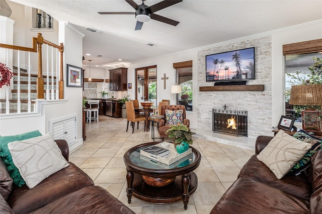 living room featuring ornamental molding, a textured ceiling, ceiling fan, a stone fireplace, and light tile patterned flooring