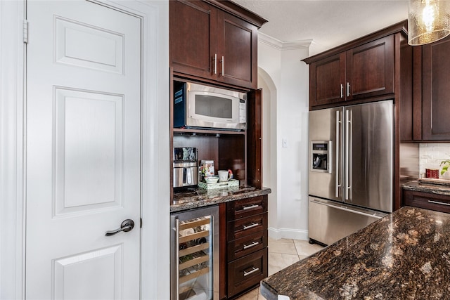 kitchen featuring dark brown cabinetry, beverage cooler, stainless steel appliances, dark stone countertops, and crown molding