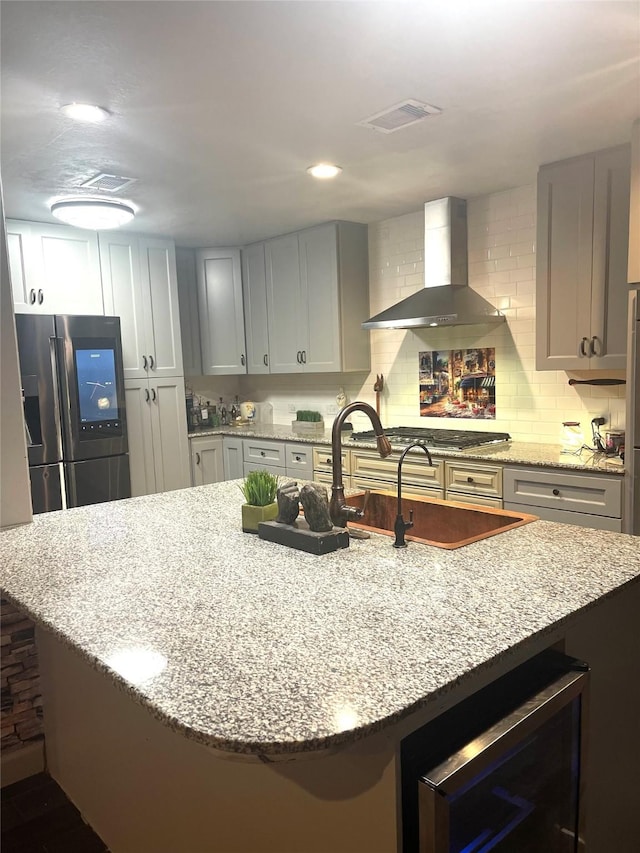 kitchen featuring a center island with sink, stainless steel appliances, wine cooler, light stone countertops, and wall chimney range hood