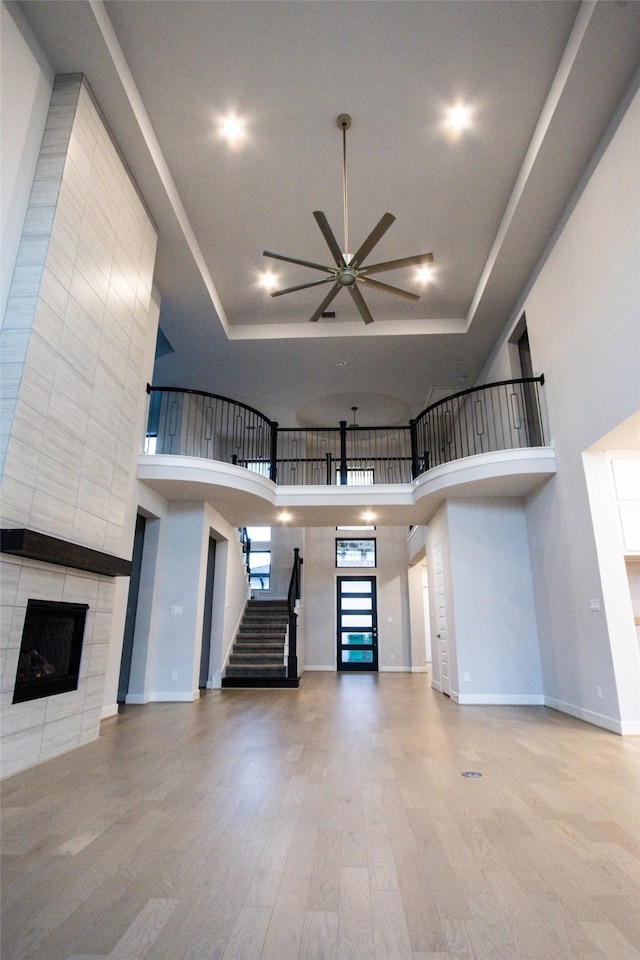 unfurnished living room with ceiling fan, a towering ceiling, a tray ceiling, a tiled fireplace, and hardwood / wood-style flooring