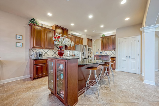 kitchen featuring backsplash, sink, stainless steel refrigerator with ice dispenser, an island with sink, and decorative columns