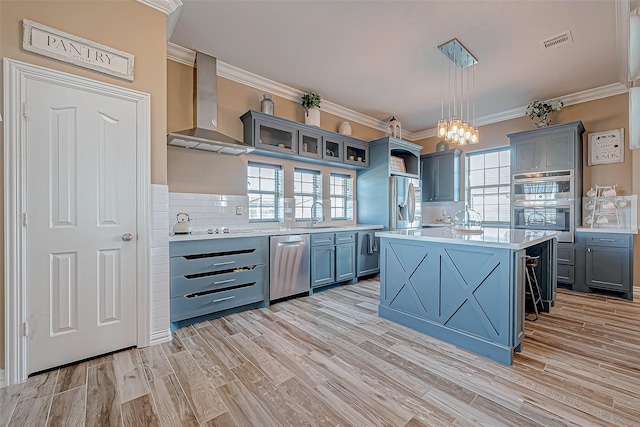 kitchen featuring wall chimney exhaust hood, stainless steel appliances, backsplash, pendant lighting, and a kitchen island with sink