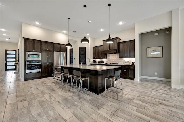 kitchen featuring dark brown cabinetry, stainless steel appliances, a center island with sink, light hardwood / wood-style flooring, and hanging light fixtures