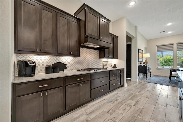 kitchen featuring light stone countertops, backsplash, dark brown cabinetry, stainless steel gas cooktop, and light hardwood / wood-style floors