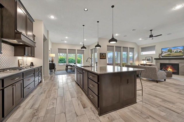 kitchen with a kitchen island with sink, a tile fireplace, decorative backsplash, ceiling fan, and stainless steel gas cooktop