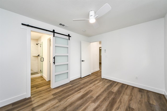 unfurnished bedroom with dark hardwood / wood-style flooring, a barn door, connected bathroom, and ceiling fan