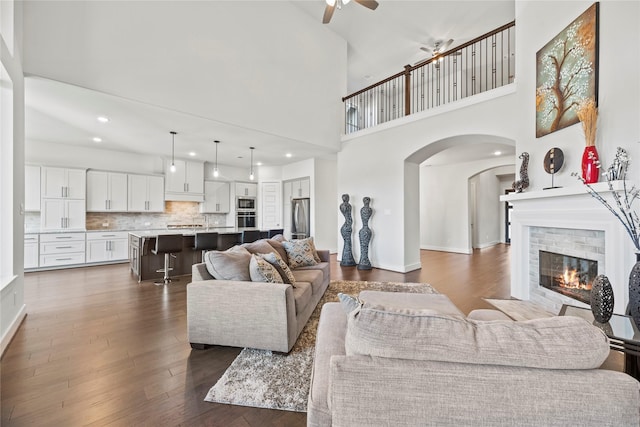 living room featuring ceiling fan, a fireplace, a towering ceiling, and dark wood-type flooring