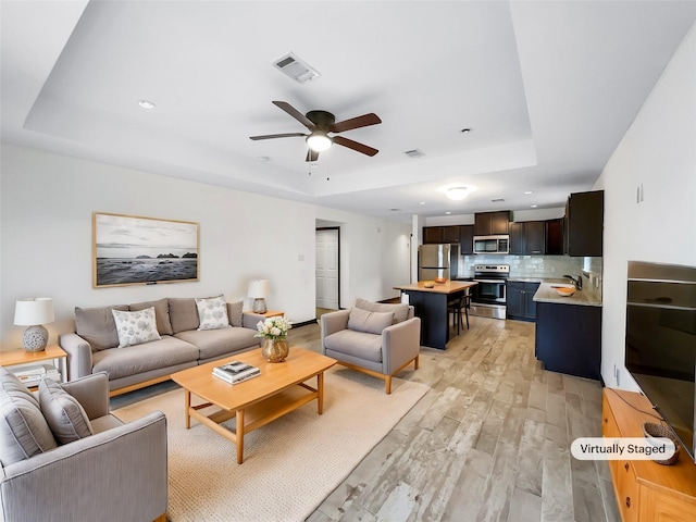 living room featuring a raised ceiling, ceiling fan, sink, and light wood-type flooring