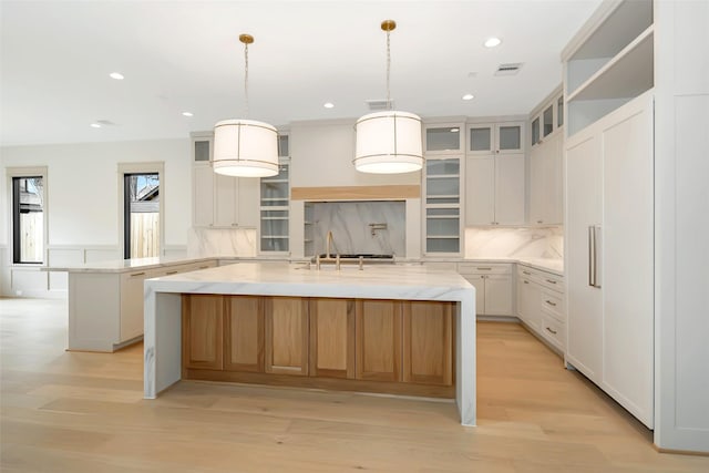 kitchen featuring light wood-type flooring, visible vents, a kitchen island with sink, tasteful backsplash, and glass insert cabinets