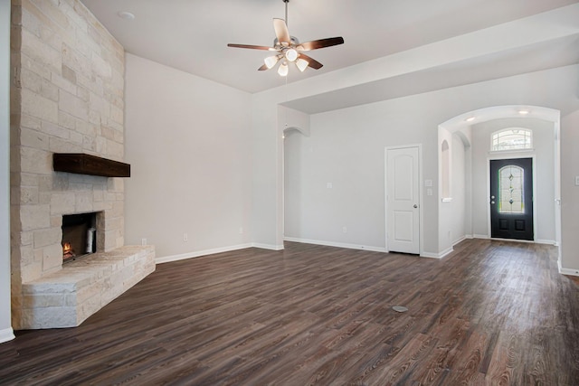 unfurnished living room with ceiling fan, a stone fireplace, and dark wood-type flooring