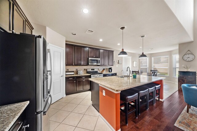 kitchen with appliances with stainless steel finishes, light stone counters, dark brown cabinetry, a kitchen island with sink, and a breakfast bar area