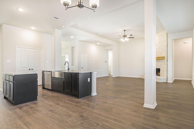 kitchen with ceiling fan with notable chandelier, a fireplace, a kitchen island with sink, and dark wood-type flooring