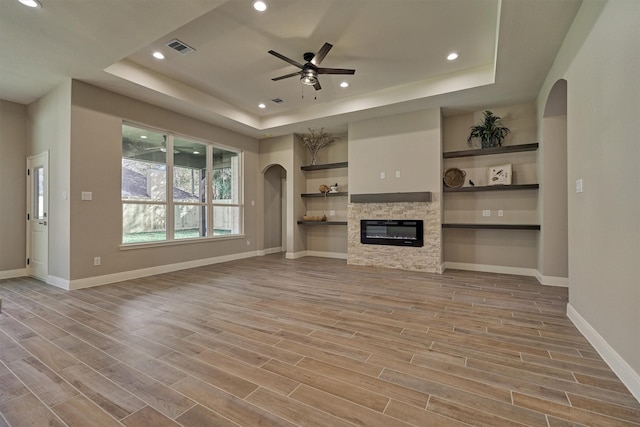 unfurnished living room with built in shelves, ceiling fan, a raised ceiling, a stone fireplace, and light hardwood / wood-style floors