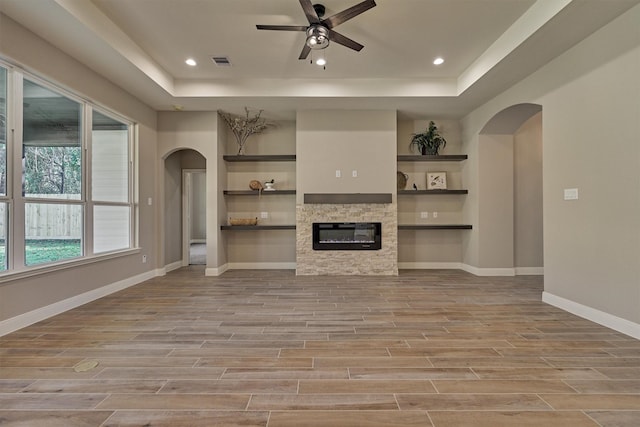 unfurnished living room with a raised ceiling, a stone fireplace, ceiling fan, and built in shelves