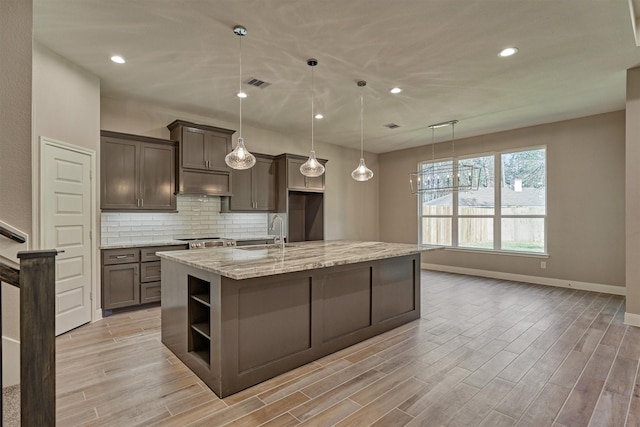 kitchen with backsplash, light stone counters, a kitchen island with sink, decorative light fixtures, and range