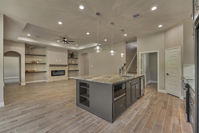 kitchen featuring pendant lighting, a tray ceiling, a stone fireplace, and a kitchen island with sink