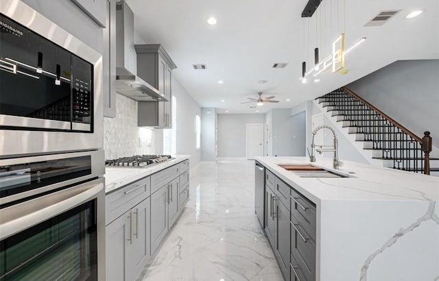 kitchen with gray cabinetry, sink, hanging light fixtures, wall chimney exhaust hood, and appliances with stainless steel finishes