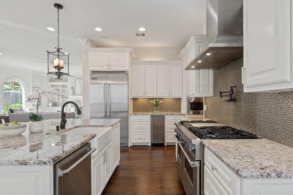 kitchen featuring light stone countertops, wall chimney range hood, pendant lighting, built in appliances, and white cabinetry