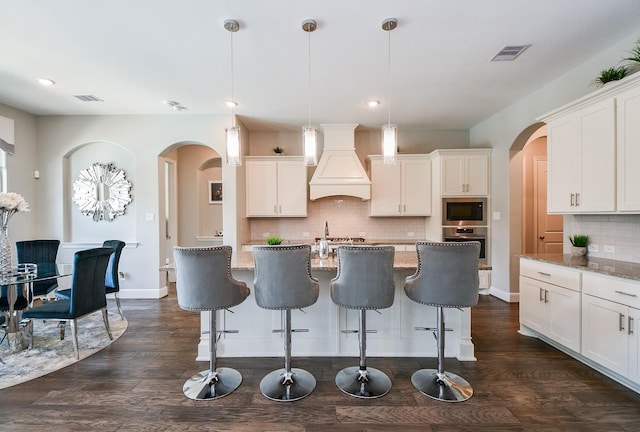 kitchen featuring custom range hood, oven, hanging light fixtures, and a kitchen island with sink