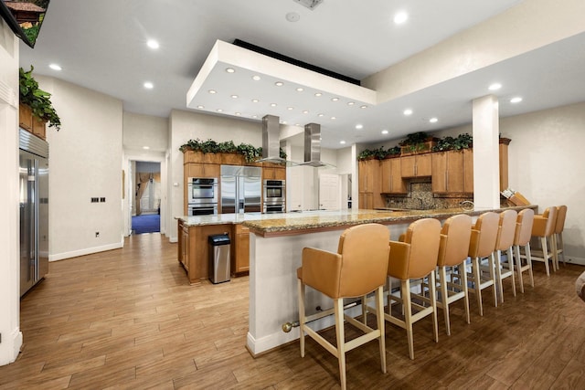 kitchen featuring wall chimney exhaust hood, kitchen peninsula, island range hood, a breakfast bar, and appliances with stainless steel finishes