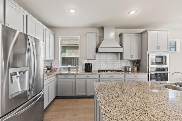 kitchen with light stone countertops, appliances with stainless steel finishes, custom exhaust hood, sink, and hardwood / wood-style flooring