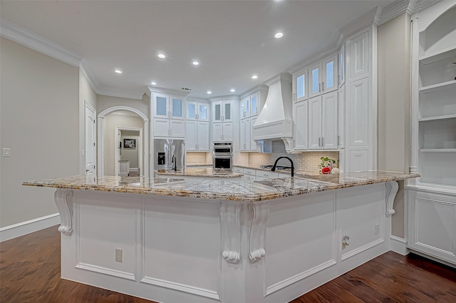 kitchen featuring stainless steel appliances, premium range hood, light stone counters, decorative backsplash, and white cabinets