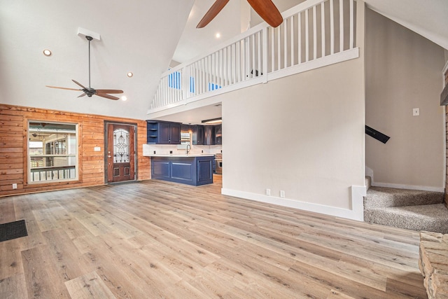 unfurnished living room with ceiling fan, a towering ceiling, wooden walls, and light hardwood / wood-style flooring