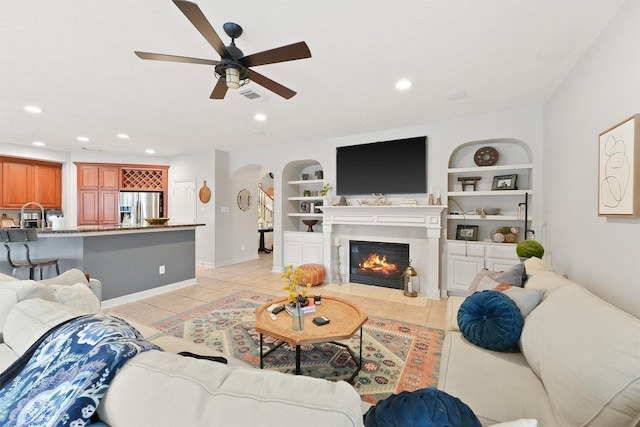 tiled living room featuring built in shelves, ceiling fan, and a tile fireplace