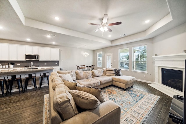 living room featuring a raised ceiling, a tiled fireplace, dark wood-type flooring, and ceiling fan with notable chandelier