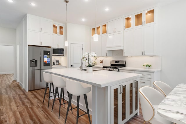 kitchen featuring appliances with stainless steel finishes, custom range hood, a kitchen island with sink, white cabinetry, and hanging light fixtures