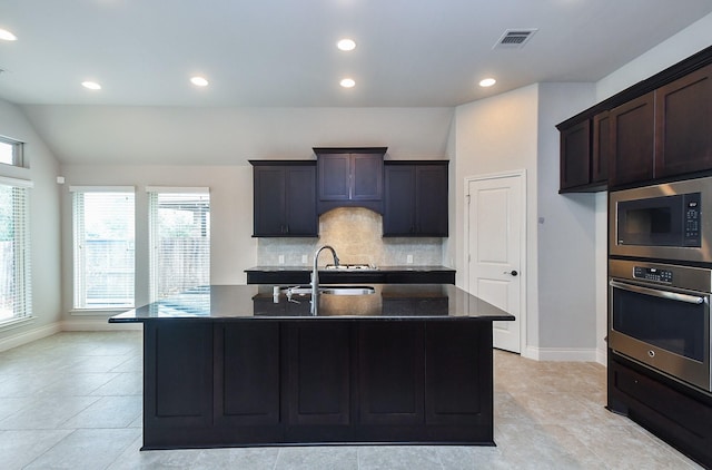 kitchen with built in microwave, sink, tasteful backsplash, stainless steel oven, and an island with sink