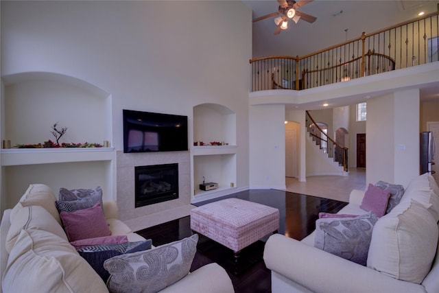 living room with built in features, hardwood / wood-style floors, a towering ceiling, a tiled fireplace, and ceiling fan