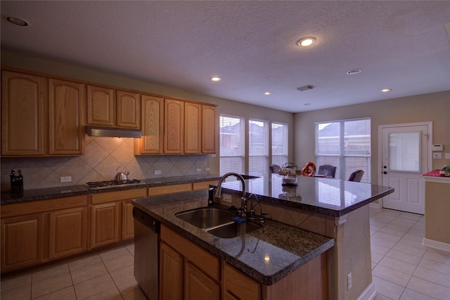 kitchen with a kitchen island with sink, appliances with stainless steel finishes, light tile patterned floors, dark stone counters, and sink