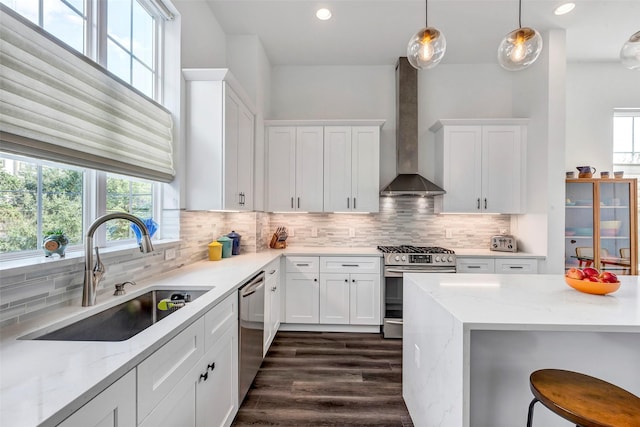 kitchen with sink, wall chimney exhaust hood, stainless steel appliances, pendant lighting, and white cabinets