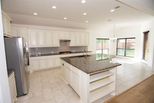 kitchen featuring stainless steel refrigerator with ice dispenser, dark stone counters, gas stovetop, a kitchen island with sink, and decorative light fixtures