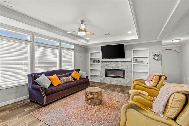 living room with ceiling fan, built in features, crown molding, a tray ceiling, and a fireplace