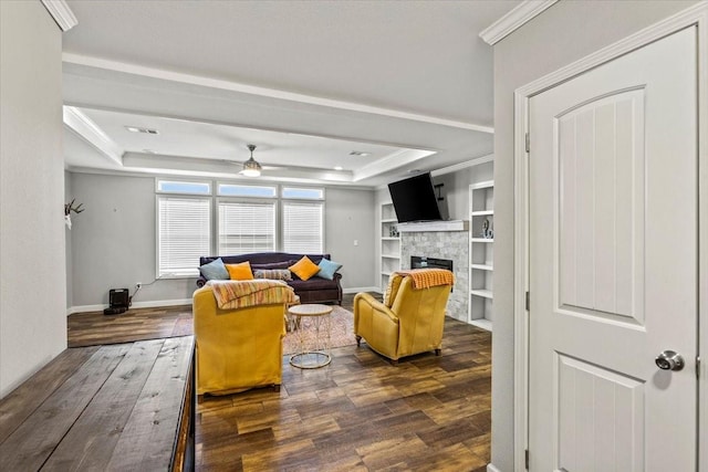 living room featuring a stone fireplace, crown molding, built in shelves, ceiling fan, and a tray ceiling