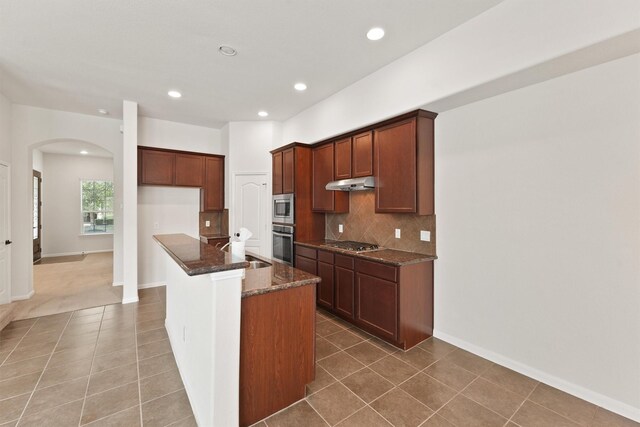 kitchen with backsplash, dark stone counters, a center island with sink, sink, and appliances with stainless steel finishes