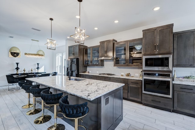 kitchen with pendant lighting, a kitchen island with sink, sink, light stone countertops, and appliances with stainless steel finishes