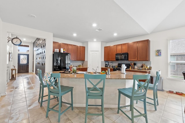 kitchen featuring a kitchen bar, light tile patterned flooring, light stone countertops, black appliances, and an island with sink