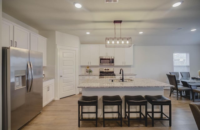 kitchen featuring appliances with stainless steel finishes, decorative light fixtures, a center island with sink, white cabinets, and light wood-type flooring