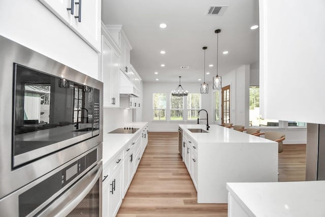 kitchen featuring black electric cooktop, sink, a center island with sink, white cabinetry, and hanging light fixtures